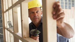 A construction worker in a hard hat uses a drill to secure a beam in a wooden frame.