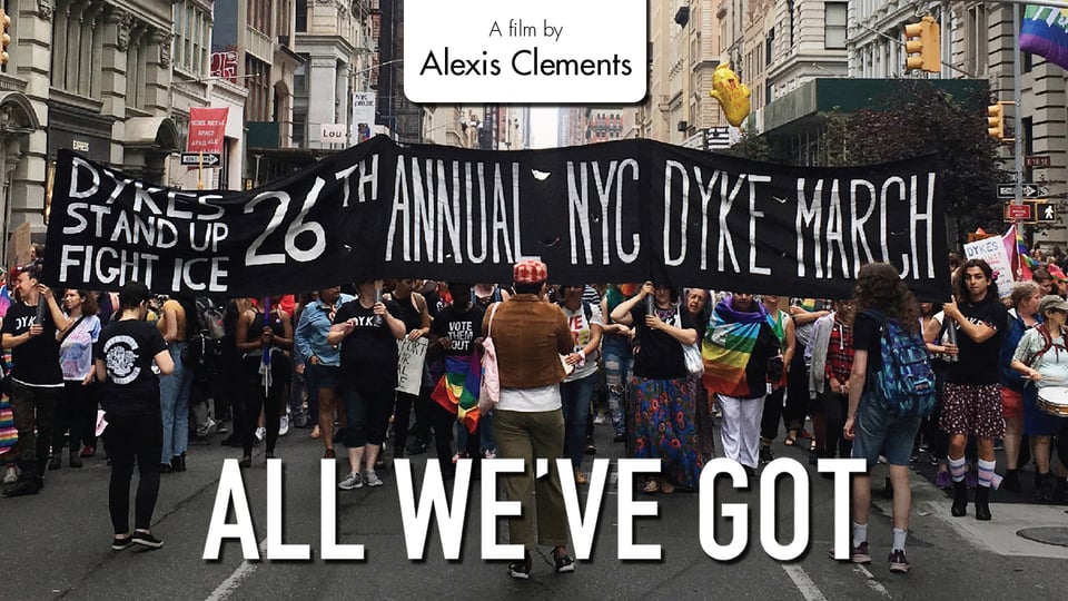 A large group of people participate in a march. They hold rainbow pride flags, a sign reading "Dyke March", and a sign reading "Dykes stand up to ICE".